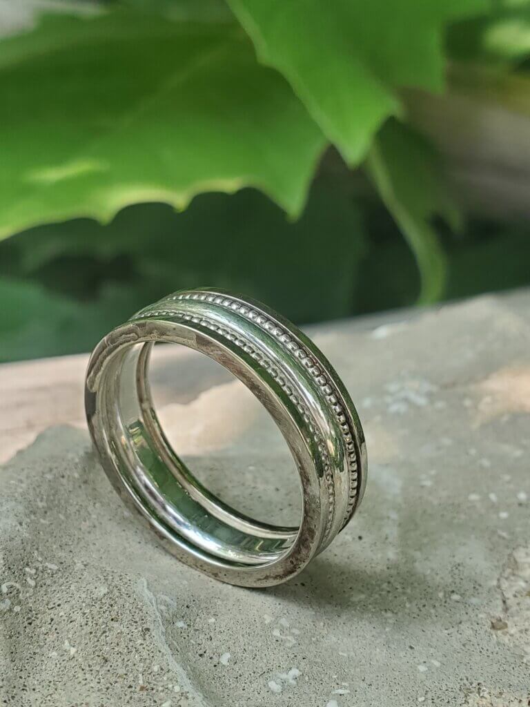 A textured sterling silver wedding band sits on a rock outside, with trees in the blurred out background. 