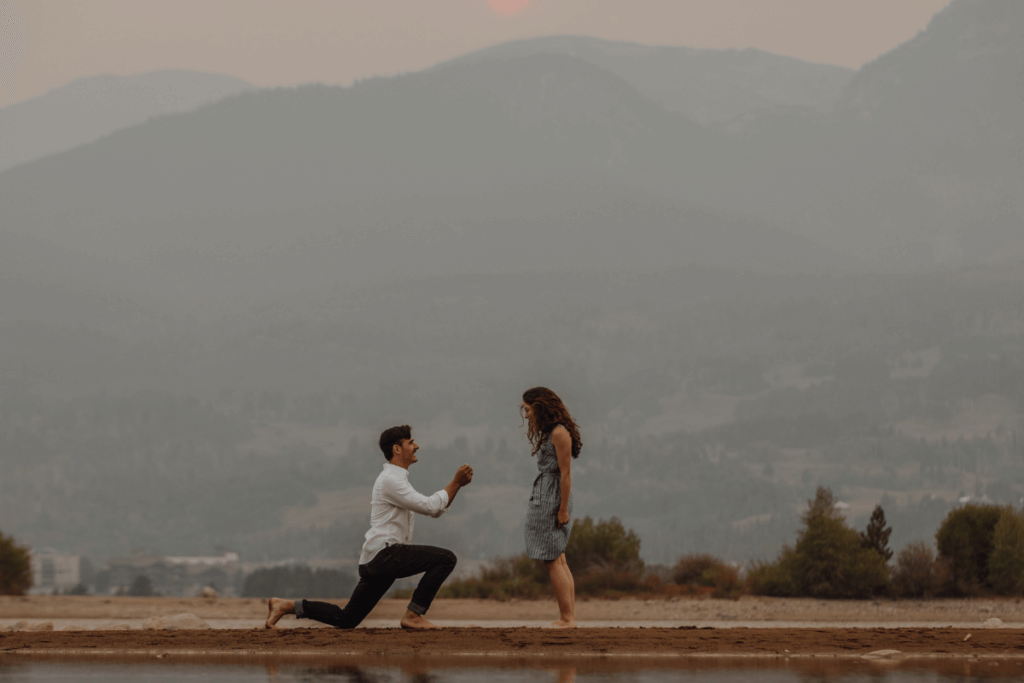 A proposal in the mountains takes place as a couple is shown smiling at each other with the man on one knee.